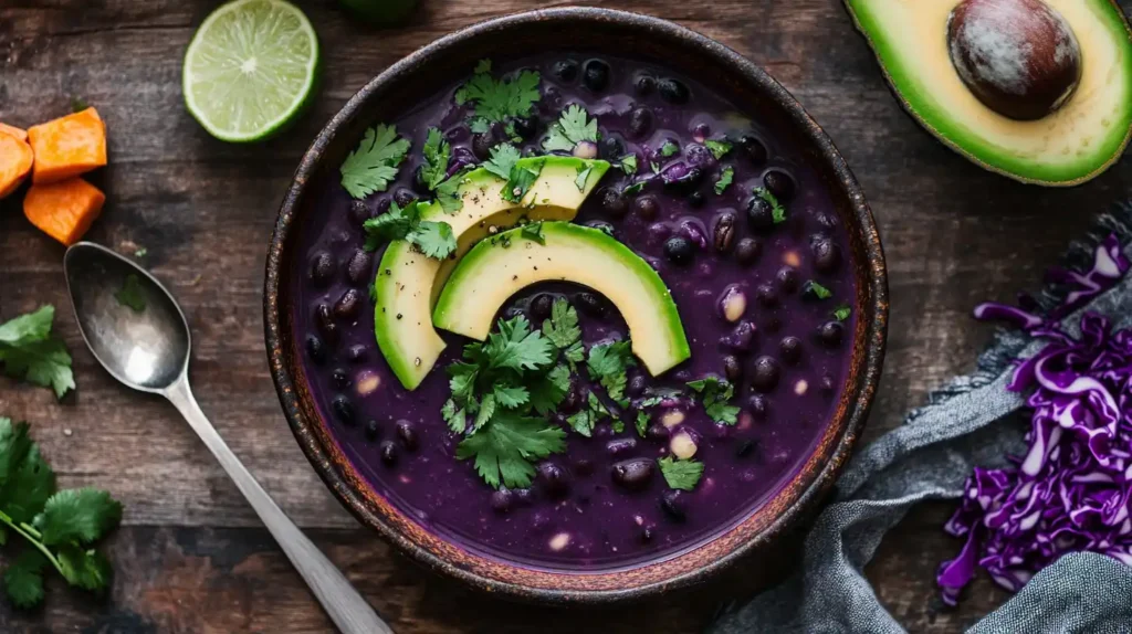 Purple black bean soup served in a rustic bowl, garnished with avocado, lime, and cilantro, with vibrant ingredients nearby.