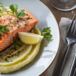 Perfectly cooked salmon fillet with creamy mashed potatoes garnished with parsley and lemon on a white plate.